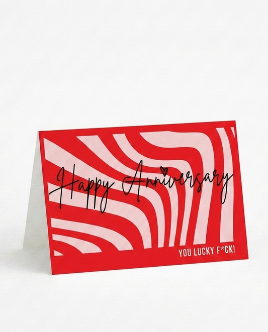 A red and white patterned standing greeting card with the words 'Happy Anniversary' in black script and 'YOU LUCKY F!' in small white text at the bottom right, photographed on a clean white background.