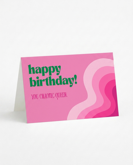 A pink birthday card standing upright on a white surface. The card has green text reading 'happy birthday!' and smaller pink text reading 'YOU CHAOTIC QUEEN.