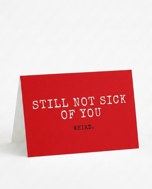 A red A5-style desk card with white typewriter-style text reading 'STILL NOT SICK OF YOU' and smaller text 'WEIRD.' below, standing upright on a white background.
