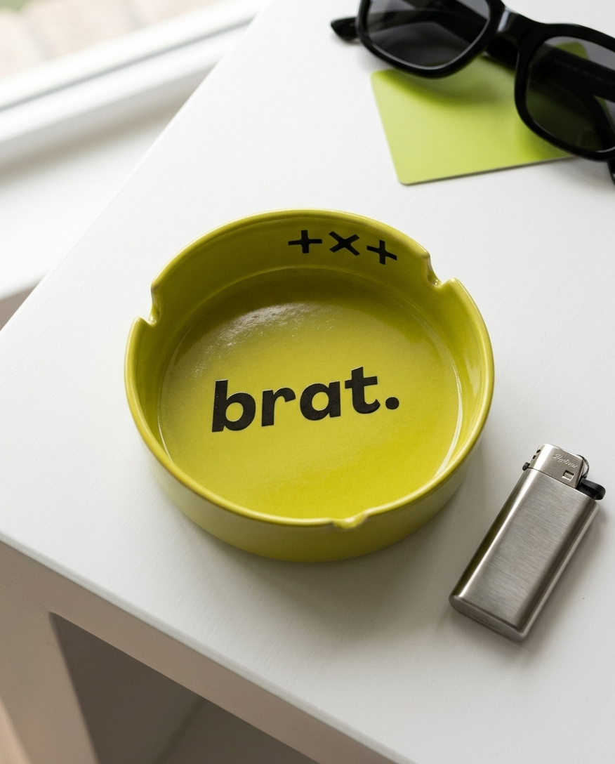 A bright yellow-green ceramic ashtray with the word 'brat.' printed in black inside, styled on a white table next to a silver lighter, black sunglasses, and a lime green sticky note.