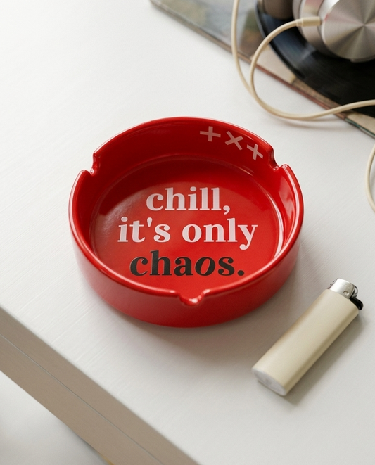 A red ceramic ashtray with the phrase 'chill, it's only chaos.' printed in white and black text, styled on a white tabletop next to a cream-colored lighter and headphones.