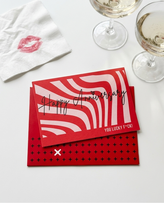Red and white anniversary greeting card stacked on a white surface, styled with two glasses of white wine, a white napkin with a pink lip print, and a red envelope