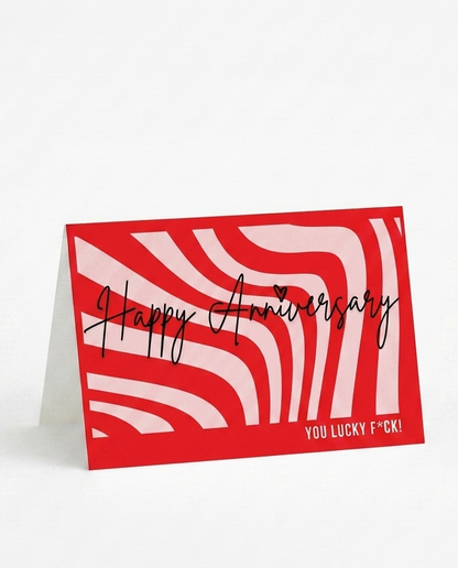 A red and white patterned standing greeting card with the words 'Happy Anniversary' in black script and 'YOU LUCKY F!' in small white text at the bottom right, photographed on a clean white background.