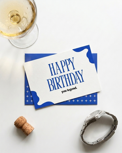 A styled flat-lay of a blue and white birthday card that reads 'HAPPY BIRTHDAY you legend.' on a white surface, with a wine glass, cork, and silver watch nearby as props.