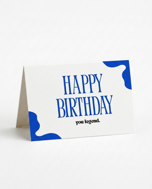 A folded white greeting card standing on a white surface, with bold blue abstract corner shapes and the text 'HAPPY BIRTHDAY you legend.' printed on it.
