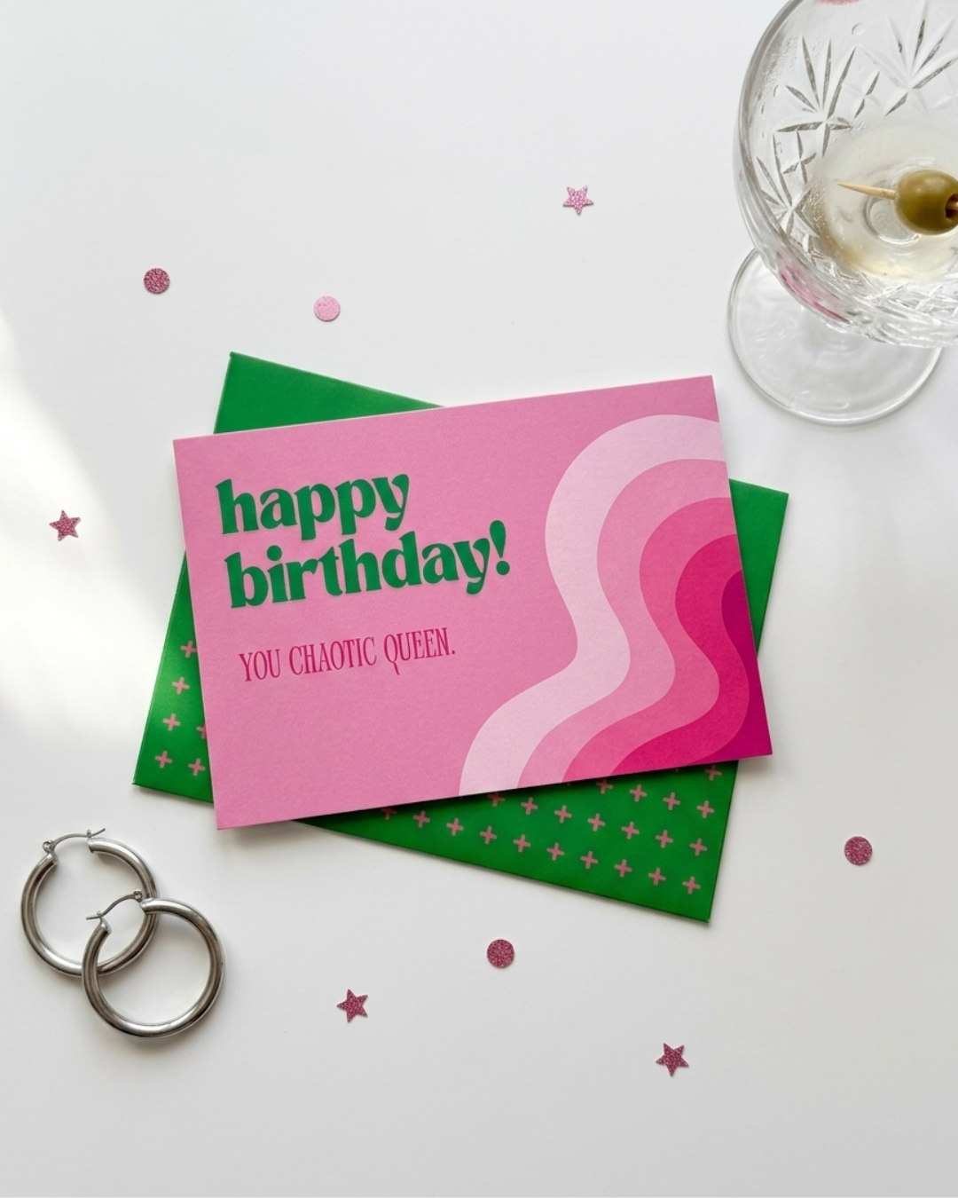 A pink and green birthday card with the text 'happy birthday! YOU CHAOTIC QUEEN.' is styled on a white surface with a green envelope, silver hoop earrings, a glass with an olive, and pink star confetti.