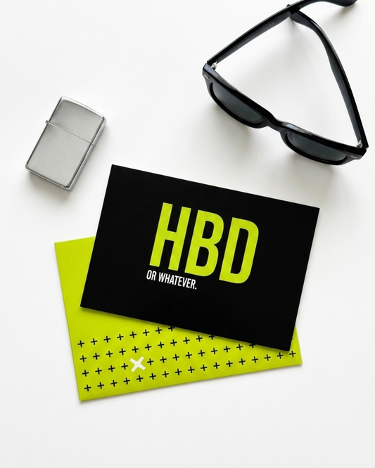 A black birthday card with bold yellow-green 'HBD OR WHATEVER.' text, placed on top of a neon lime envelope