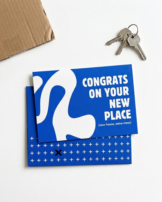 A blue and white congratulations card reading 'CONGRATS ON YOUR NEW PLACE (new house, same mess)' is stacked on top of an envelope. The scene is styled on a white surface with a brown cardboard box corner and a pair of keys nearby.