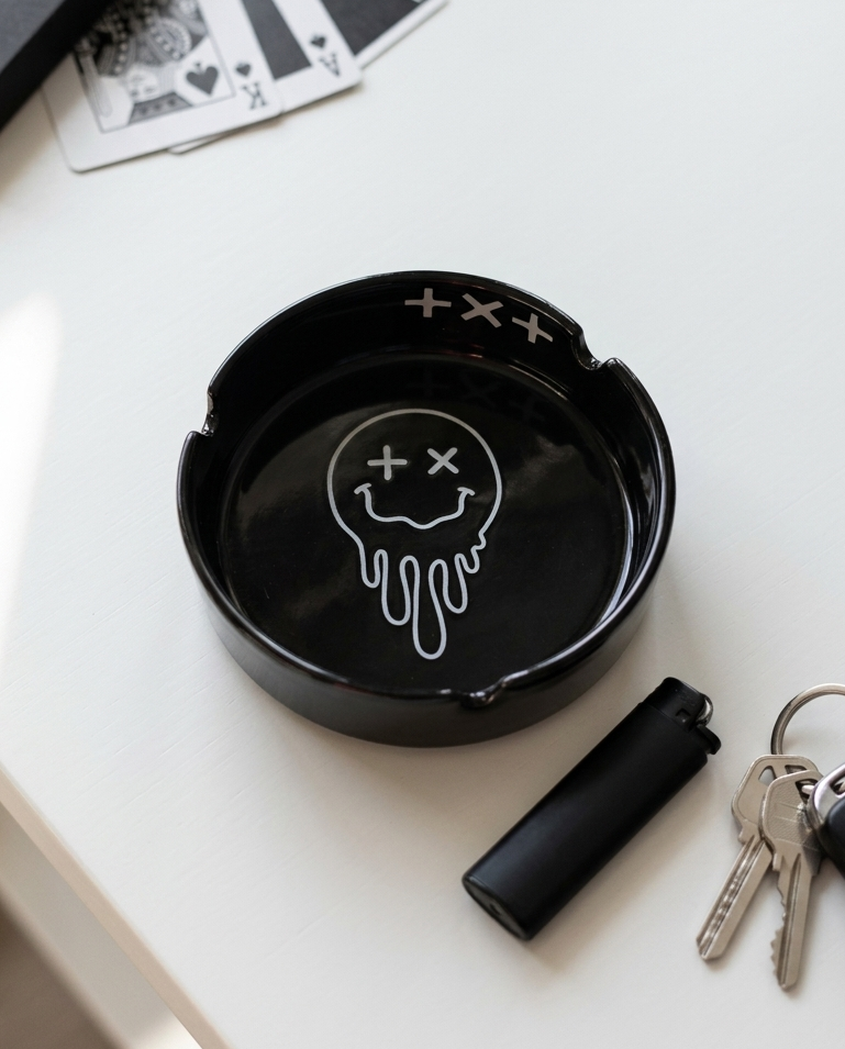 A black ceramic ashtray with a white dripping smiley face graphic, styled on a white tabletop next to keys, a lighter, and playing cards.