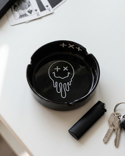 A black ceramic ashtray with a white dripping smiley face graphic, styled on a white tabletop next to keys, a lighter, and playing cards.