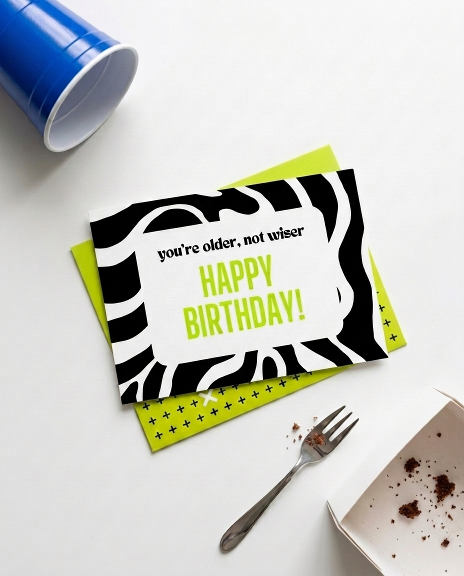A birthday card with a black and white zebra print border and lime green text reading 'you're older, not wiser HAPPY BIRTHDAY!' is displayed on a white surface with a matching lime green patterned envelope, a blue cup, a fork, and a box of cake crumbs.