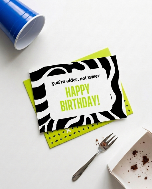 A birthday card with a black and white zebra print border and lime green text reading 'you're older, not wiser HAPPY BIRTHDAY!' is displayed on a white surface with a matching lime green patterned envelope, a blue cup, a fork, and a box of cake crumbs.