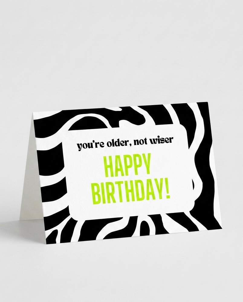 A zebra-print birthday card standing upright on a light gray background. The card has a white center panel with the text 'you're older, not wiser' in black and 'HAPPY BIRTHDAY!' in bright yellow-green.