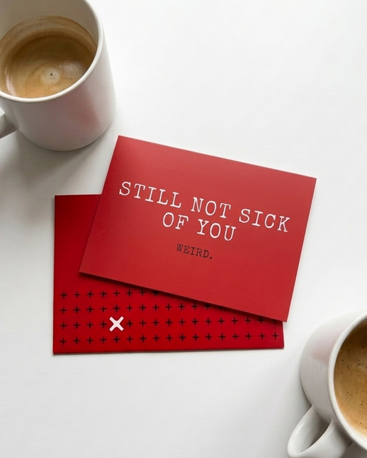 A red anniversary card with white text reading 'STILL NOT SICK OF YOU' lies on a white surface, partially covering a red envelope. 