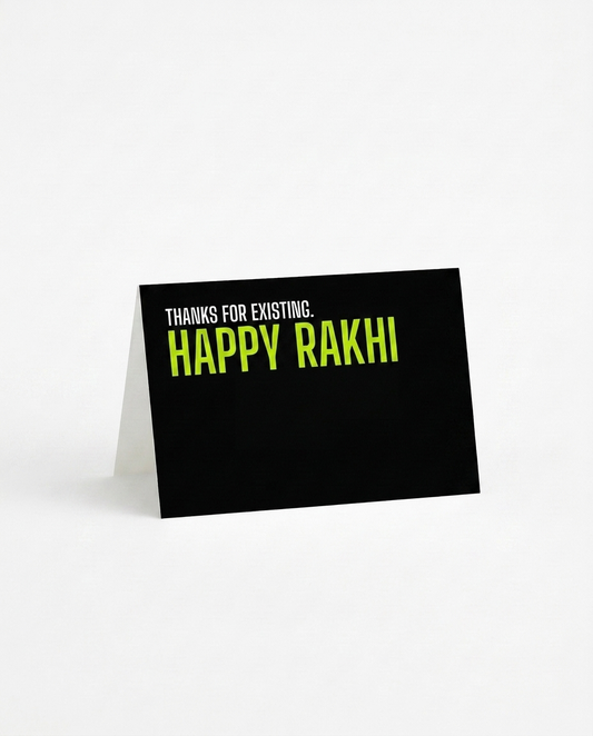 A black rakhi card with a white fold-over back, standing upright on a white surface. The front features the text 'THANKS FOR EXISTING. HAPPY RAKHI.'
