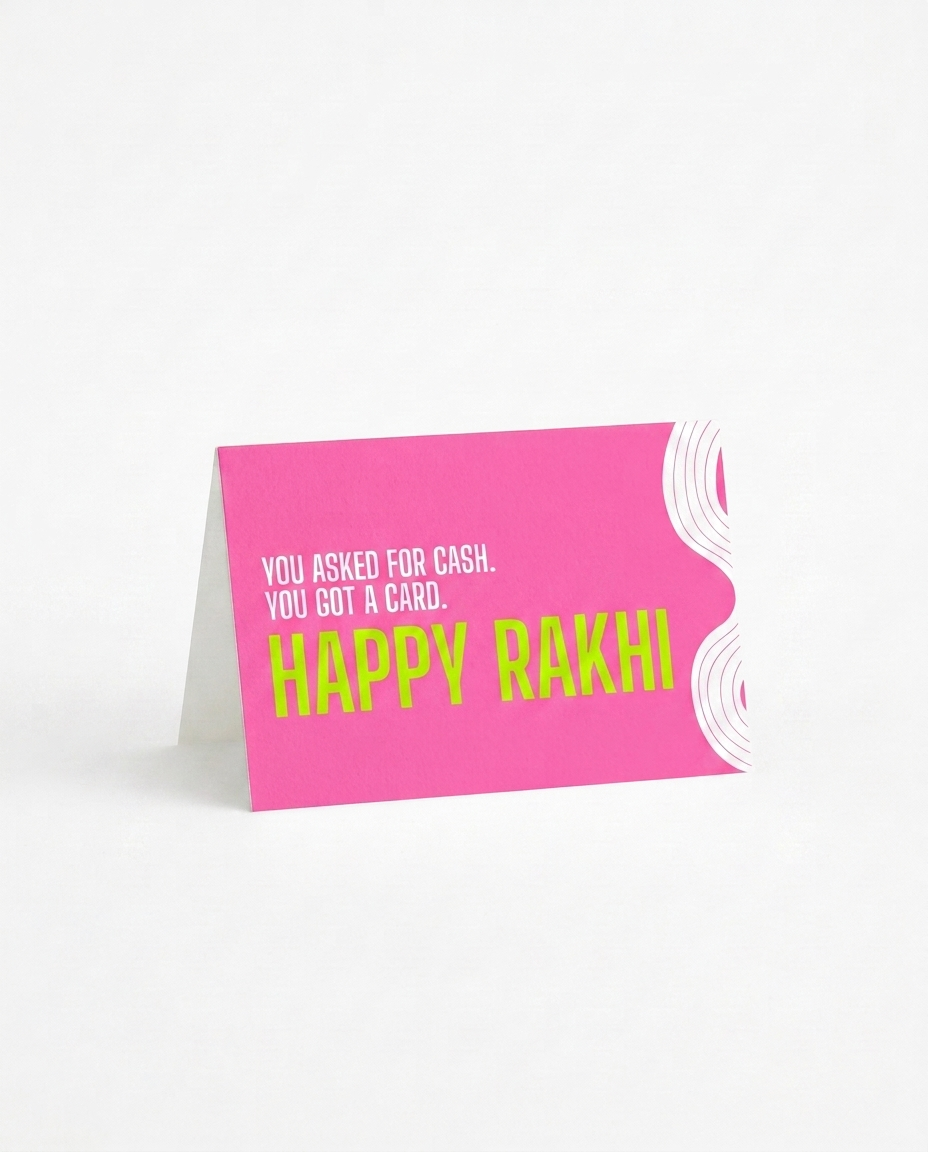 A bright pink folded greeting card standing upright on a white background. The card has white text reading 'YOU ASKED FOR CASH. happy rakhi
