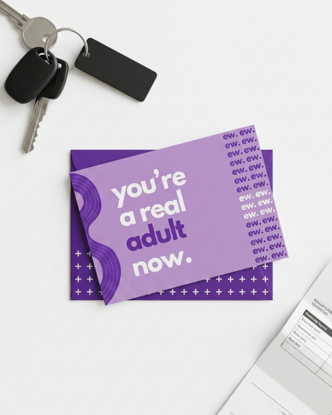 A purple adult birthday card with the text 'you're a real adult now.' and 'EW, EW, EW' printed on it, styled on a white surface with keys and papers.