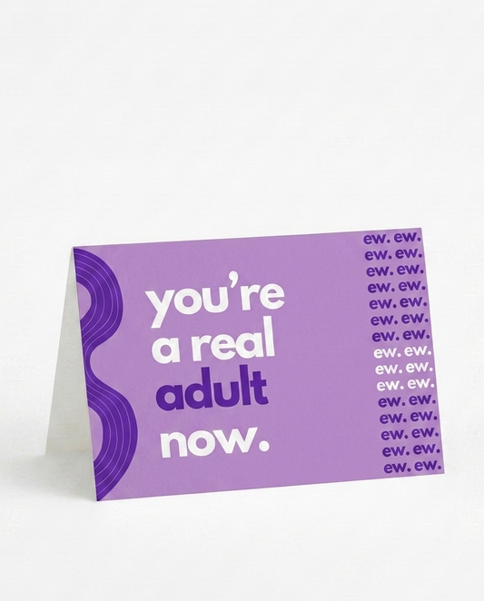 A purple greeting card standing upright on a white surface against a white background. The card features the text 'you're a real adult now.